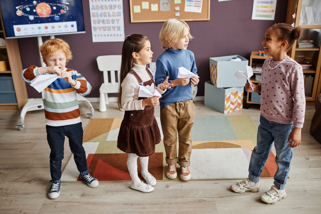 Several intercultural kids in casualwear playing paper planes and having fun while standing in classroom in nursery school after lesson
